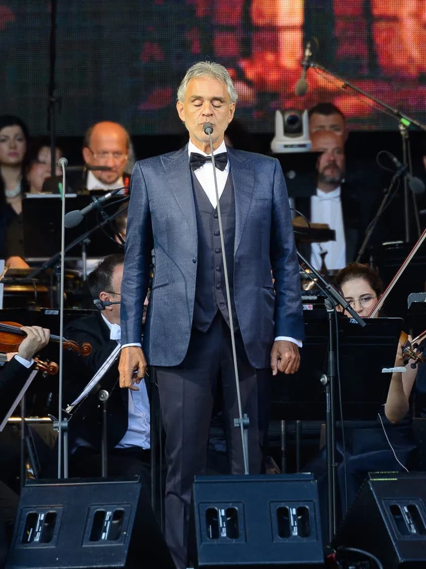 Andrea Bocelli sings into a microphone on stage, wearing a blue suit and bow tie, with an orchestra visible in the background.