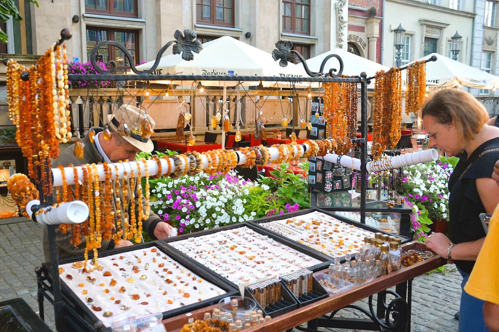 A man in a hat tends to an outdoor stall loaded with amber jewelry while a woman in a black shirt examines the items.
