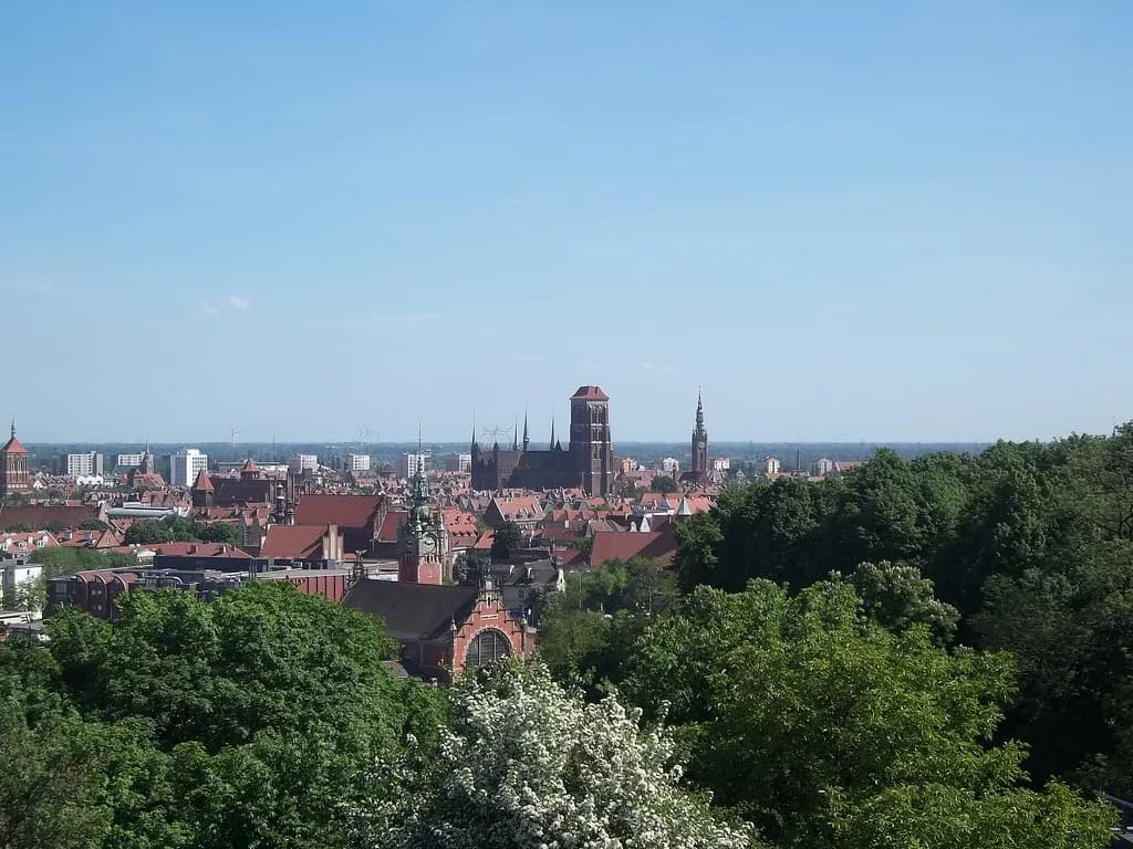 An aerial view of a sprawling European city with red-tiled roofs and several prominent church spires, surrounded by green trees under a clear blue sky.