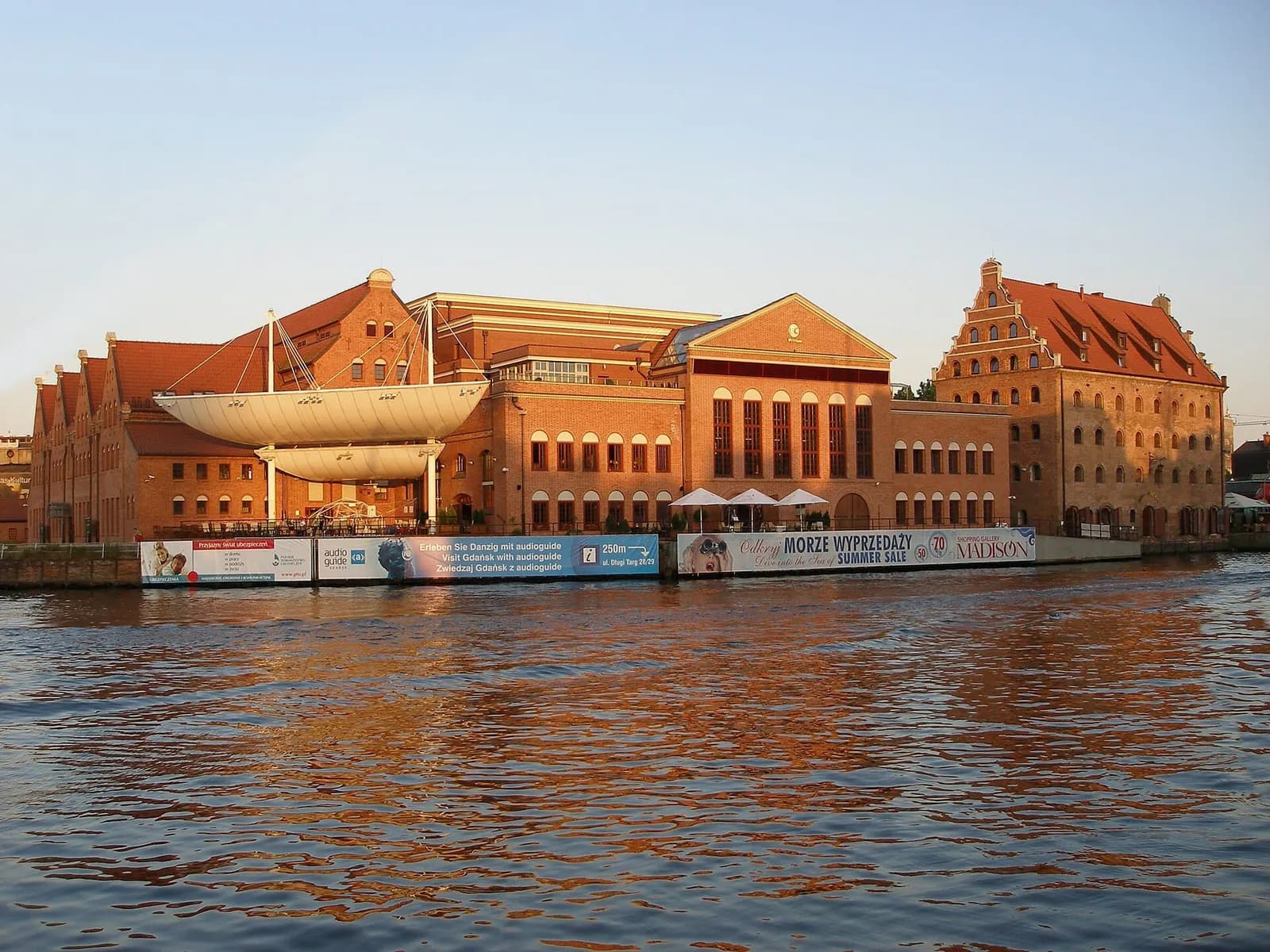 A large white boat sculpture against a backdrop of historic brick buildings with red roofs by a body of water reflecting the sunlight.